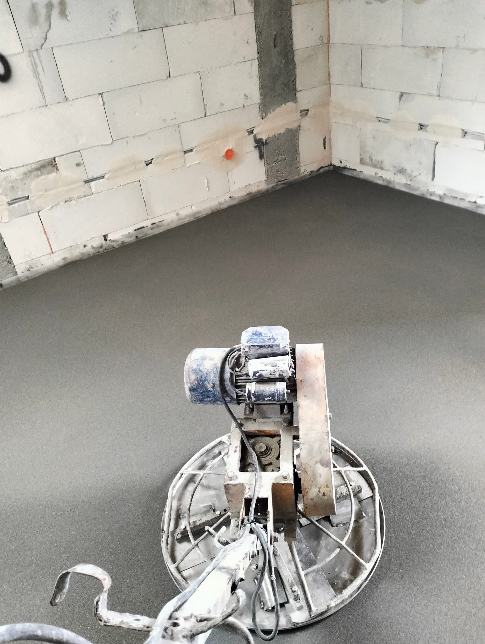 Zacieraczka mechaniczna na świeżo wylanej, gładkiej posadzce w surowym wnętrzu z białymi bloczkami. Widok z góry na maszynę i fragment podłogi.