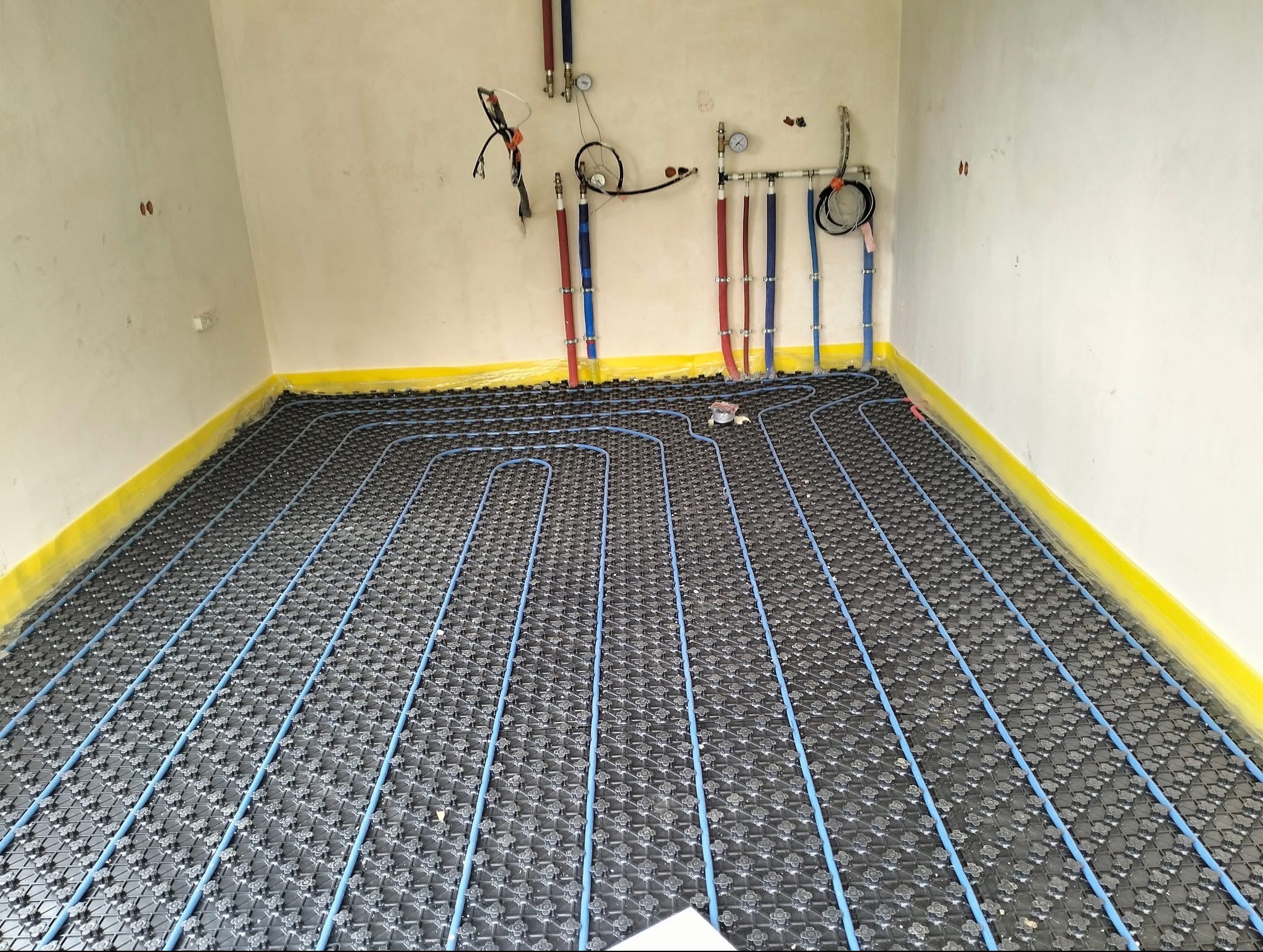 Instalacja ogrzewania podłogowego: niebieskie rurki na czarnej macie z wypustkami, żółta folia przy ścianach. Widoczne rury instalacji wodnej na ścianie.