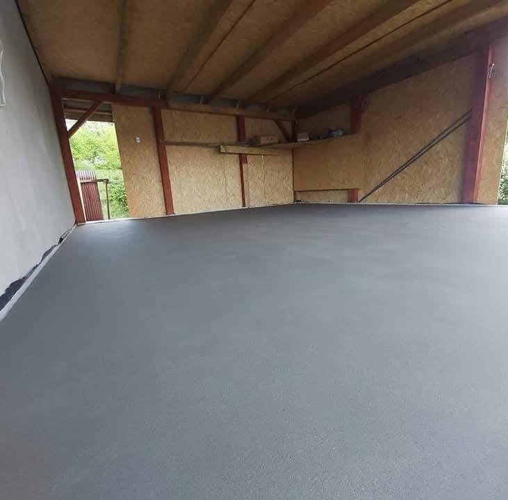 Szara, gładka posadzka w surowym wnętrzu z drewnianą konstrukcją stropu i ścian. Widoczne niedoskonałości ścian i drzwi na zewnątrz. Minimalistyczny, industrialny wygląd.