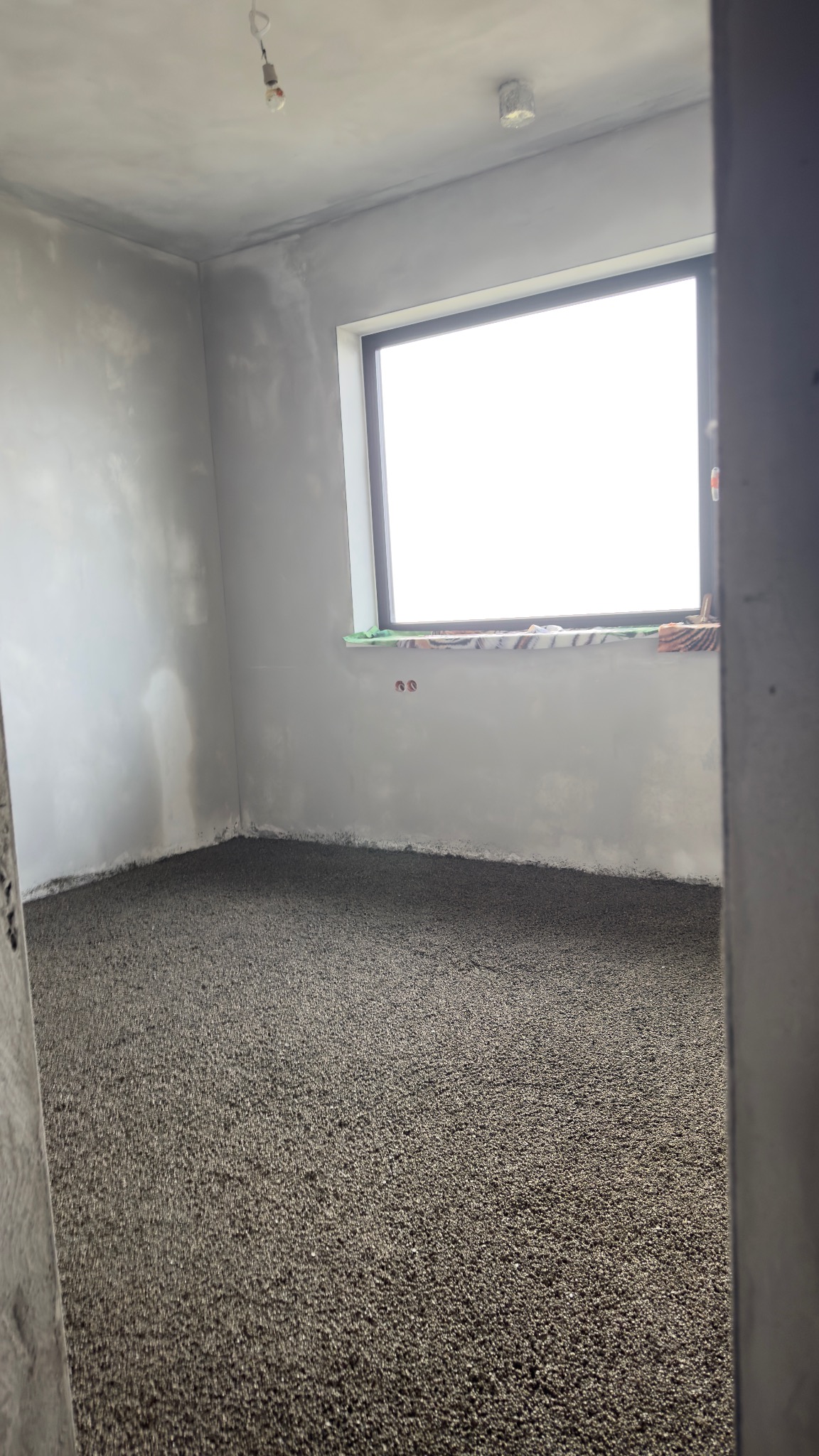 Szara, świeżo wylana posadzka w surowym wnętrzu z oknem. Widoczne ślady po łatach na ścianach. Minimalistyczne oświetlenie.