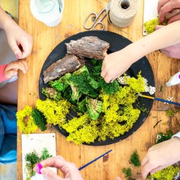 Przedsiębiorstwo Handlowo - Usługowe Beata Jasińska - Warsztaty florystyczne dla dzieci: grupa osób tworzy kompozycję z mchu i drewna na drewnianym stole. Widoczne narzędzia i materiały florystyczne. Ujęcie z góry.