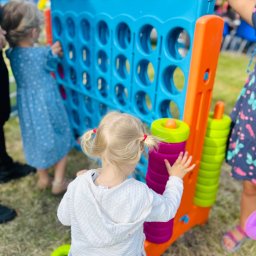 Przedsiębiorstwo Handlowo - Usługowe Beata Jasińska - Dzieci bawią się dużą, kolorową grą zręcznościową na świeżym powietrzu. Widoczne elementy gry i fragmenty ubrań dzieci. Tło rozmyte, zielona trawa.