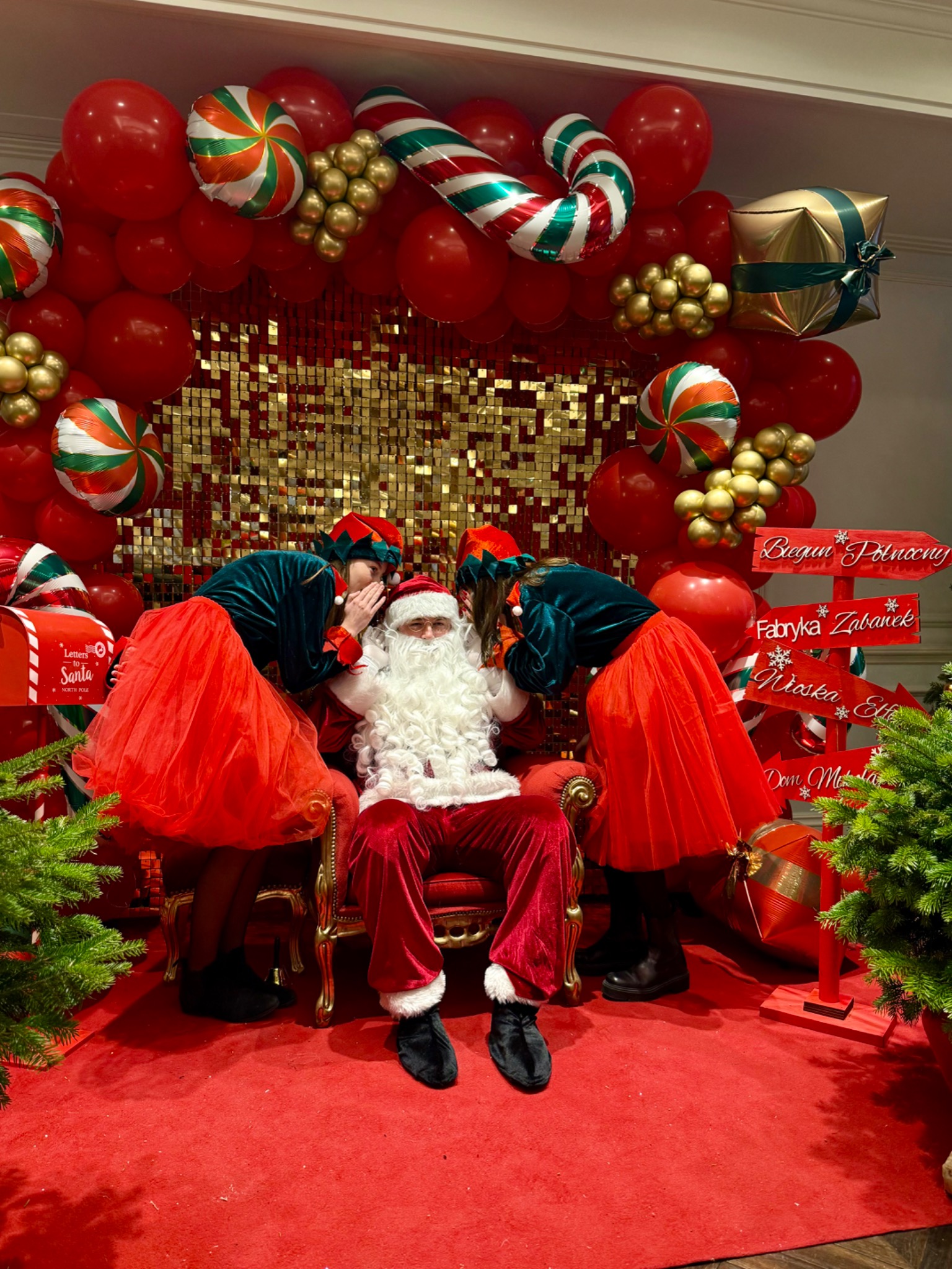 Świąteczna sesja: Mikołaj siedzi na tronie, a elfy szepczą mu do ucha na tle czerwonych balonów i złotych dekoracji. Uwagę przykuwa scenografia z drogowskazami.