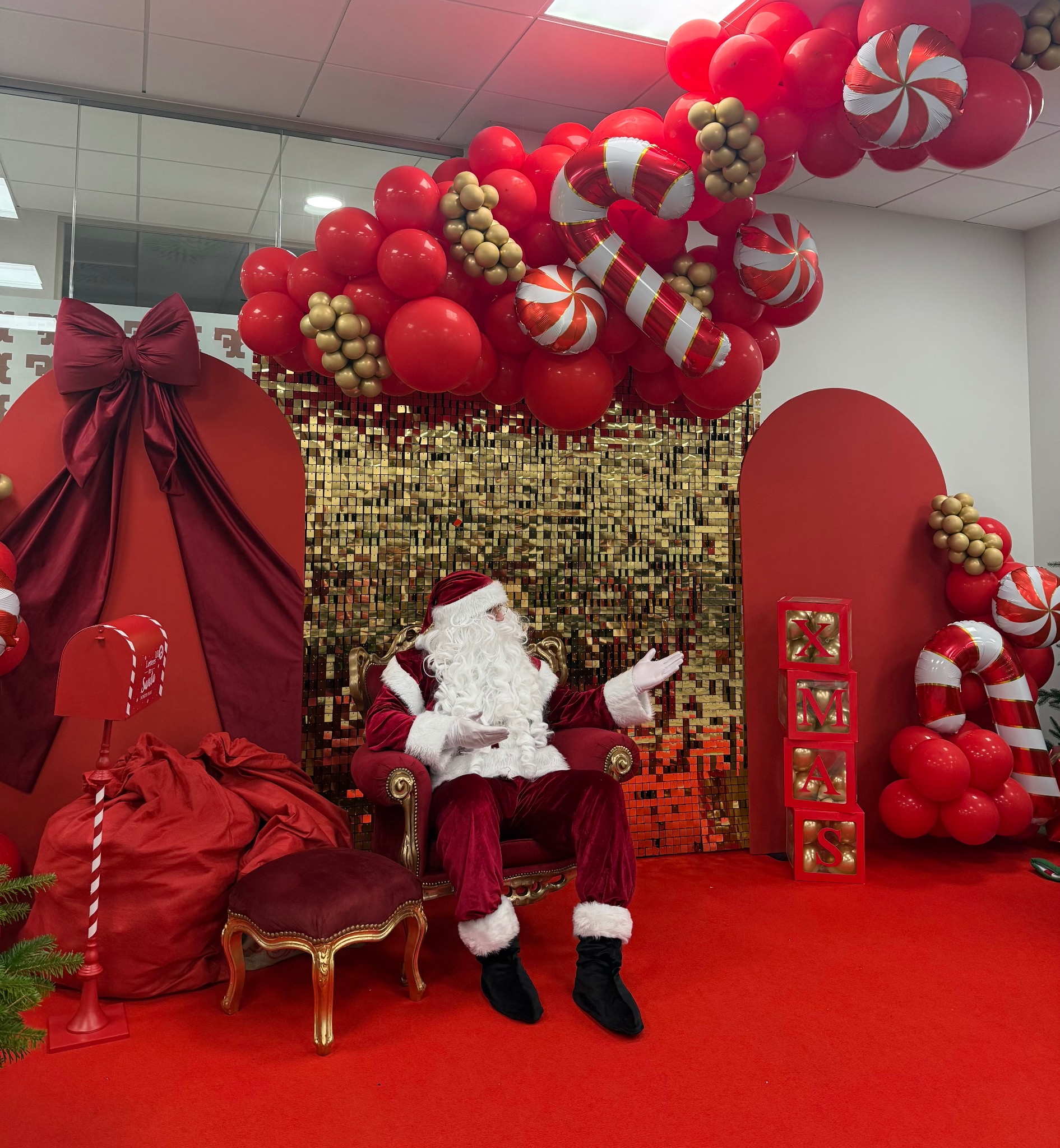 Świąteczna aranżacja z Mikołajem siedzącym na czerwonym tronie, otoczonym balonami, dekoracjami w kształcie lasek cukrowych i błyszczącym złotym tłem.