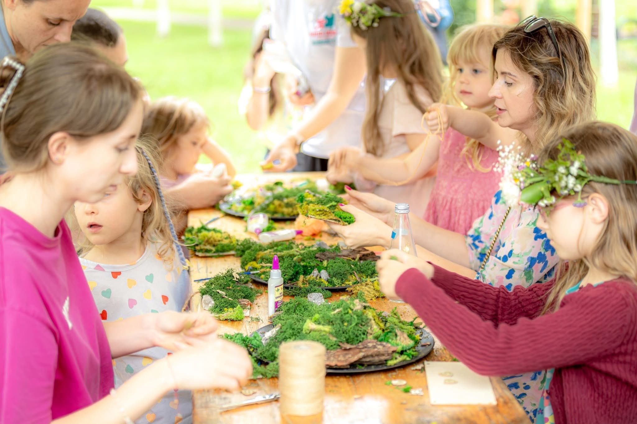 Warsztaty florystyczne dla dzieci: grupa dziewczynek i dorosłych tworzy kompozycje z mchu i kwiatów na drewnianym stole. Kreatywne zajęcie w plenerze.