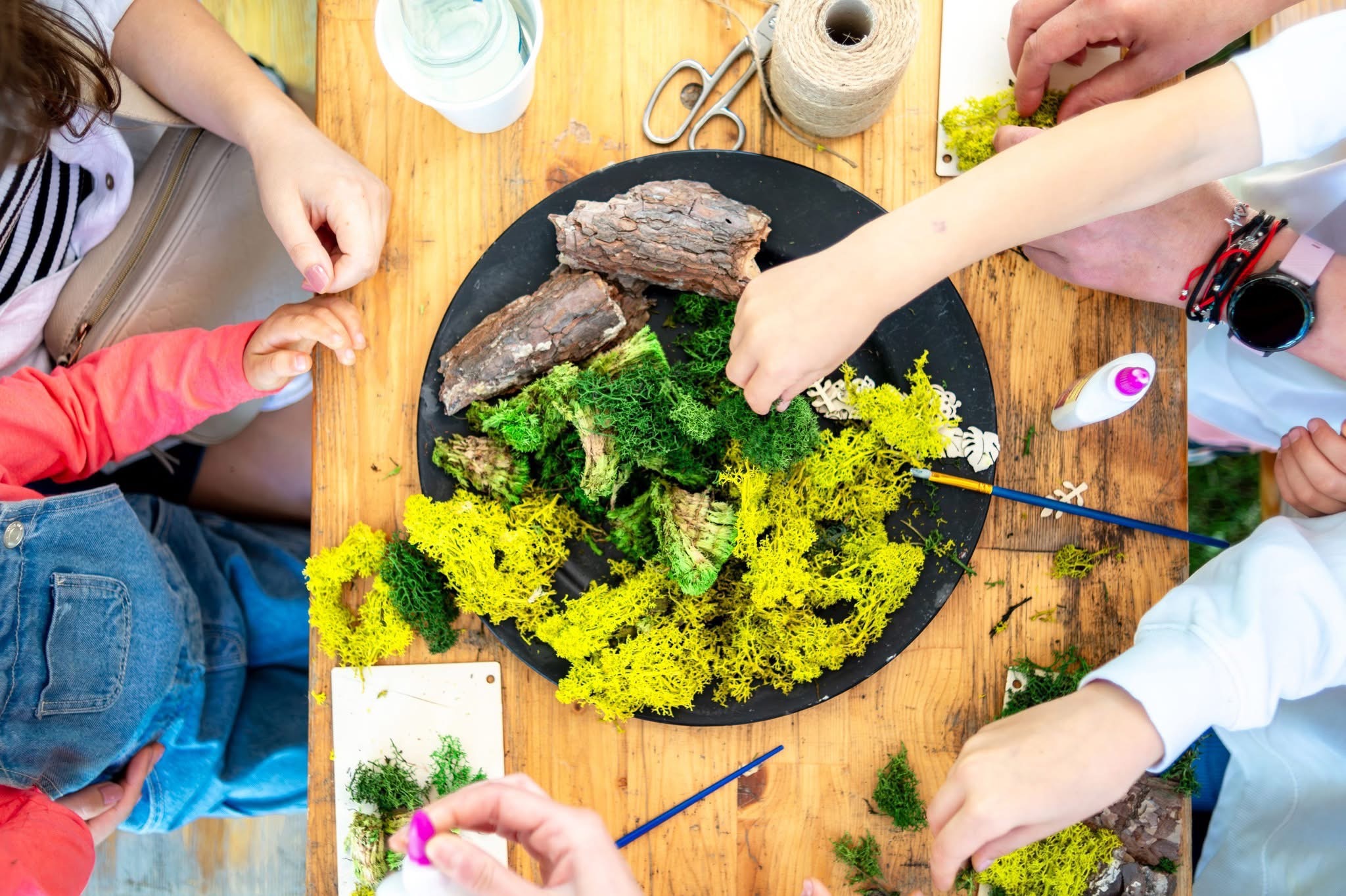Warsztaty florystyczne dla dzieci: grupa osób tworzy kompozycję z mchu i drewna na drewnianym stole. Widoczne narzędzia i materiały florystyczne. Ujęcie z góry.