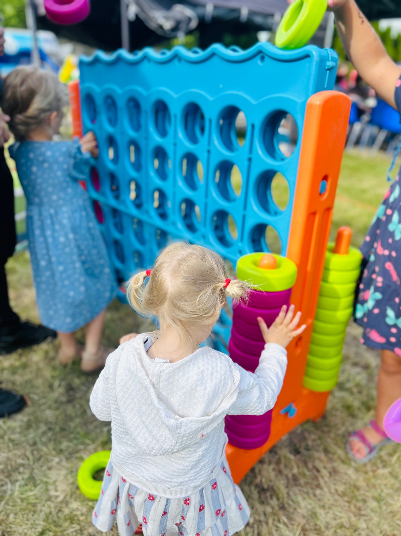 Dzieci bawią się dużą, kolorową grą zręcznościową na świeżym powietrzu. Widoczne elementy gry i fragmenty ubrań dzieci. Tło rozmyte, zielona trawa.