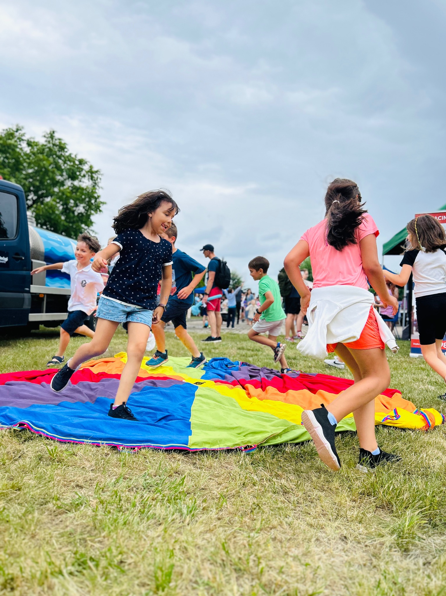 Radosne dzieci biegają po kolorowej chuście animacyjnej na trawie podczas imprezy plenerowej. W tle widoczny tłum i pojazd.