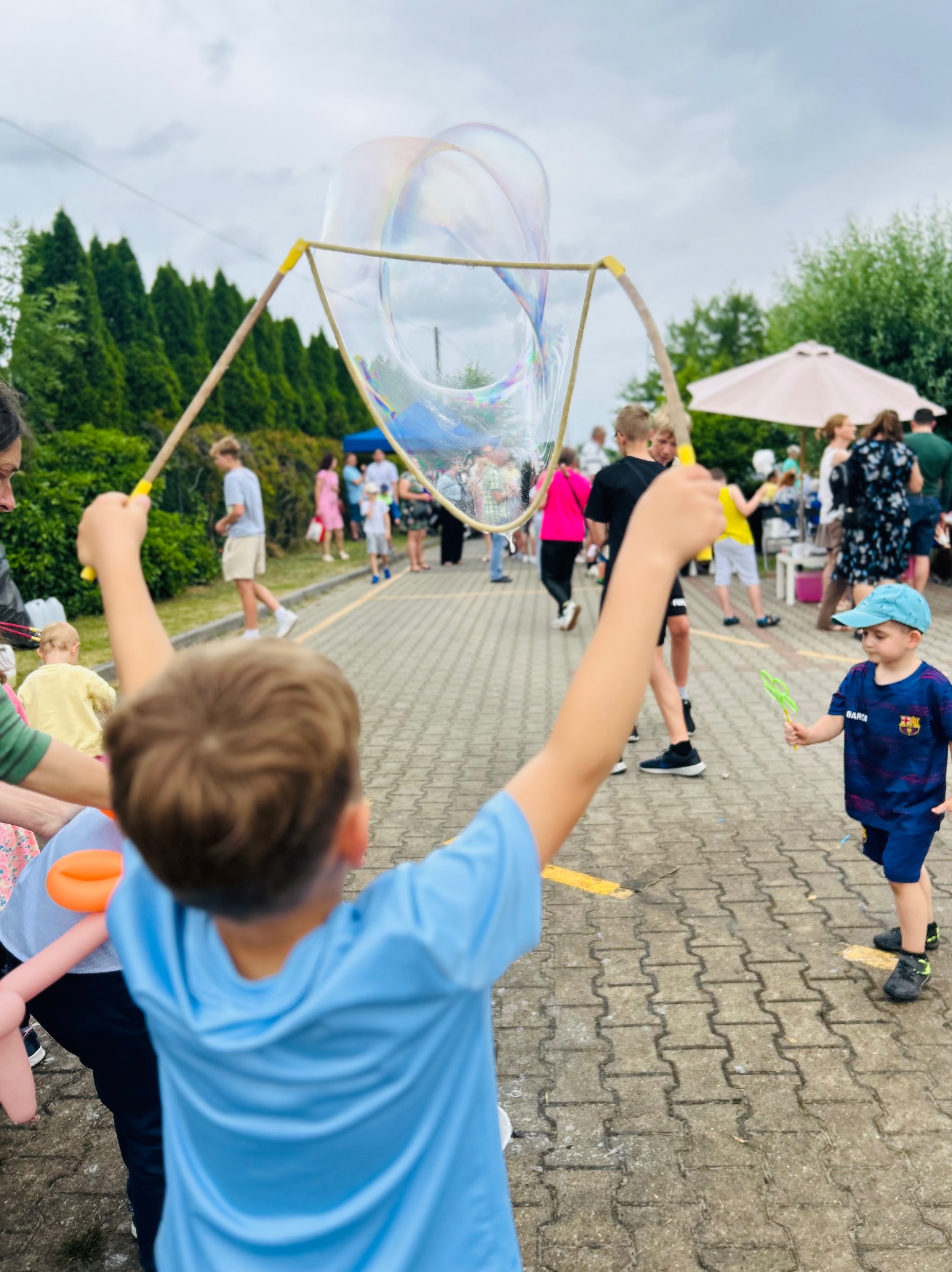 Chłopiec puszcza ogromne bańki mydlane na imprezie plenerowej, w tle widoczni inni uczestnicy zabawy i stoisko z parasolem.