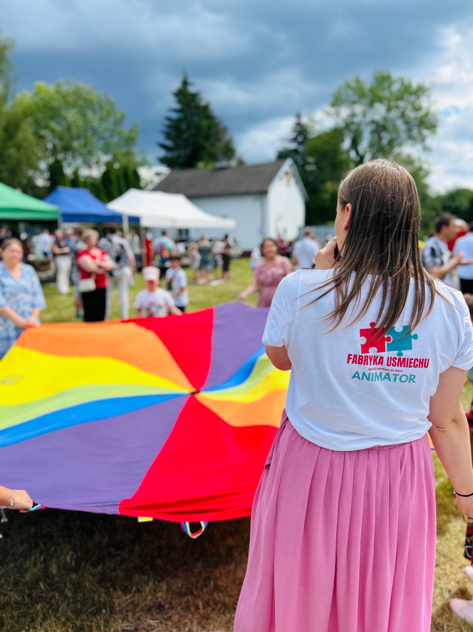Animatorka w koszulce z logo 'Fabryka Uśmiechu' prowadzi zabawy z kolorową chustą animacyjną na tle plenerowej imprezy z namiotami i uczestnikami.