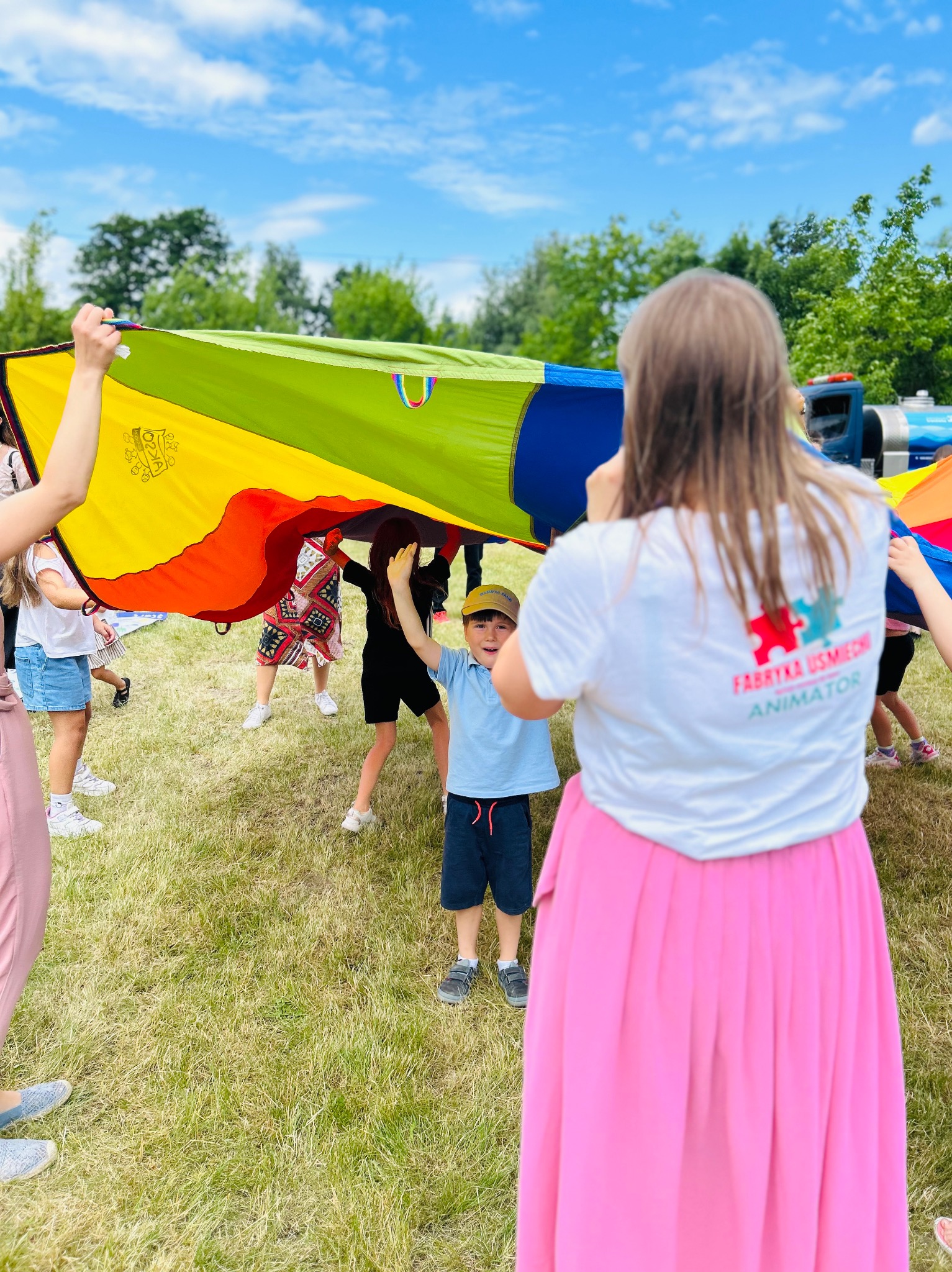 Animator w akcji: dzieci bawią się pod kolorową chustą animacyjną na trawie. Widoczna osoba z tyłu w koszulce z napisem 'Fabryka Uśmiechu Animator'.
