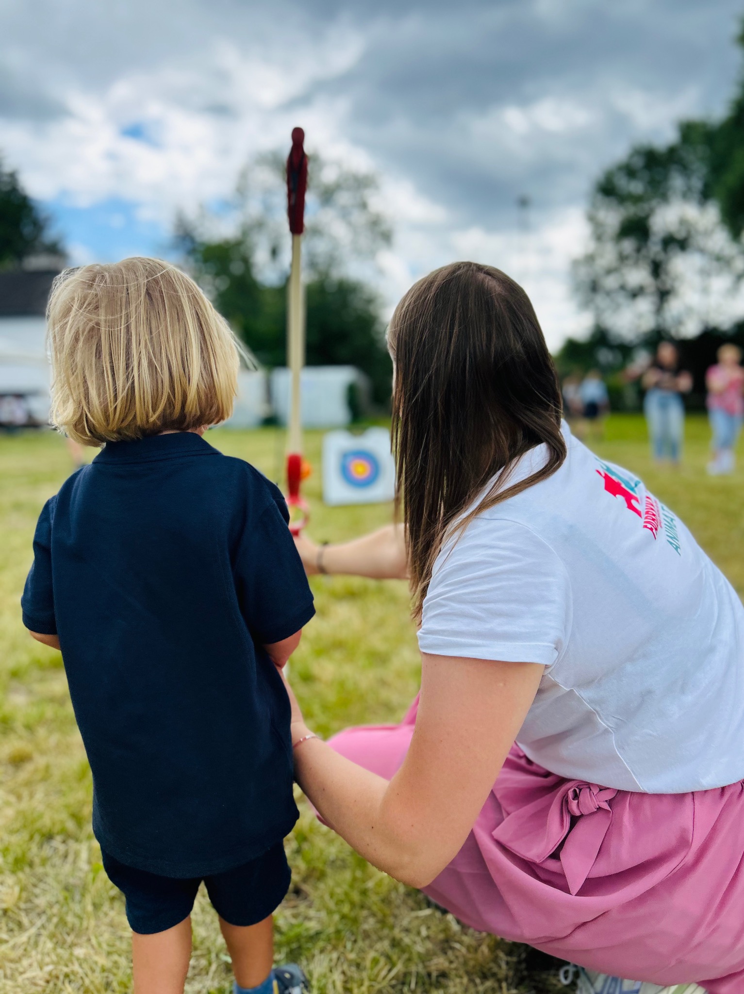 Instruktorka z dzieckiem na trawie, uczący się strzelać z łuku do tarczy. Na koszulce instruktorki logo firmy animacyjnej. Aktywność na świeżym powietrzu.