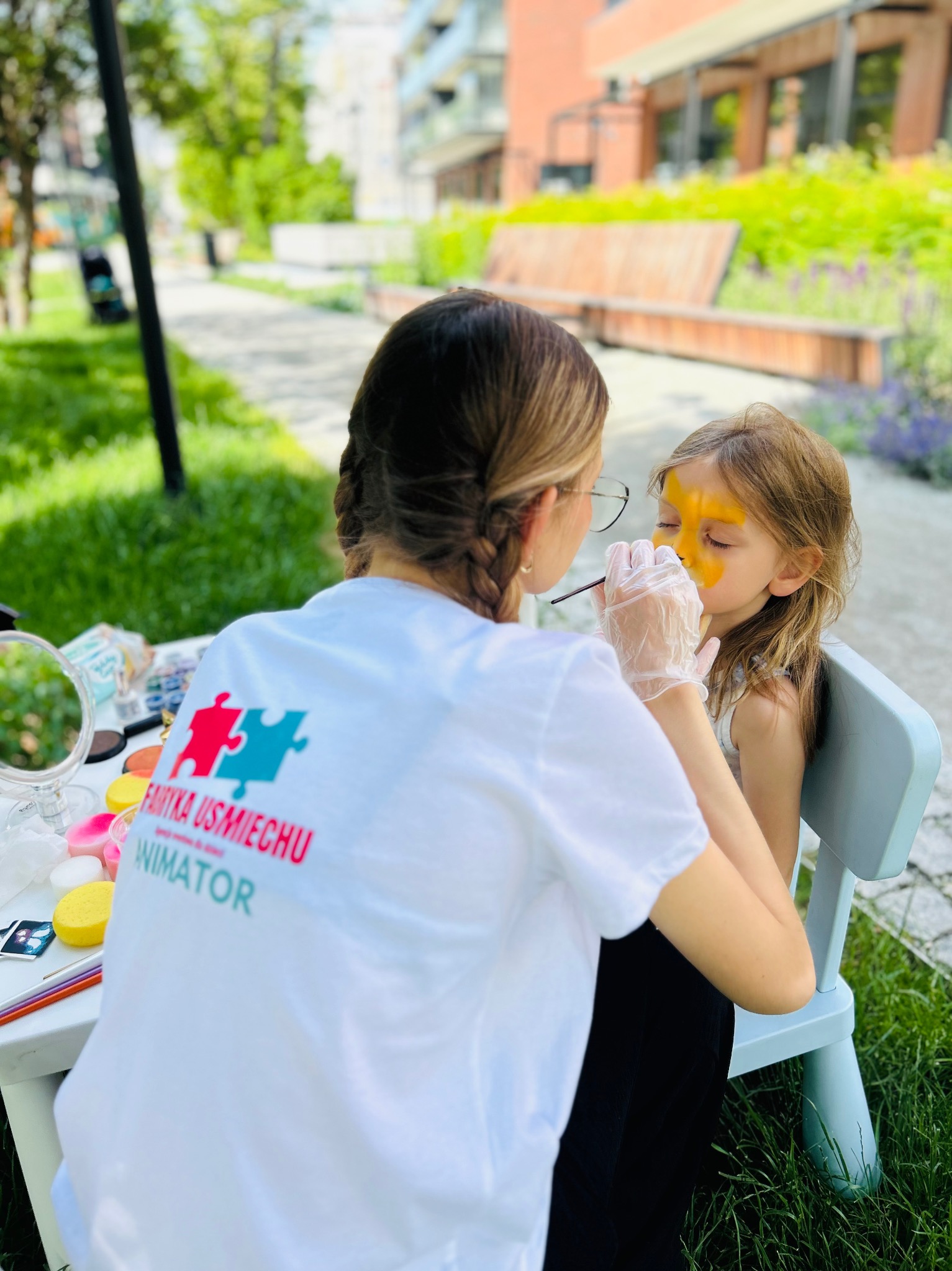 Animator maluje twarz dziewczynce w plenerze. Na stole akcesoria do malowania, w tle budynki i zieleń. Widoczny logotyp firmy animacyjnej na koszulce.