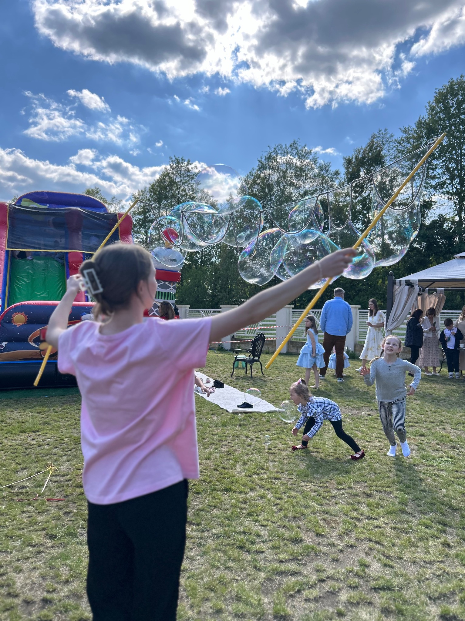 Animator puszcza gigantyczne bańki mydlane na trawie podczas imprezy plenerowej. W tle dmuchany zamek i goście.