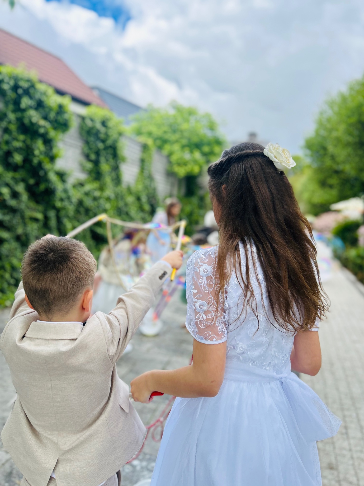 Dziewczynka w białej sukience i chłopiec w beżowym garniturze puszczają bańki mydlane na zewnątrz. Uroczystość z animatorem dla dzieci.