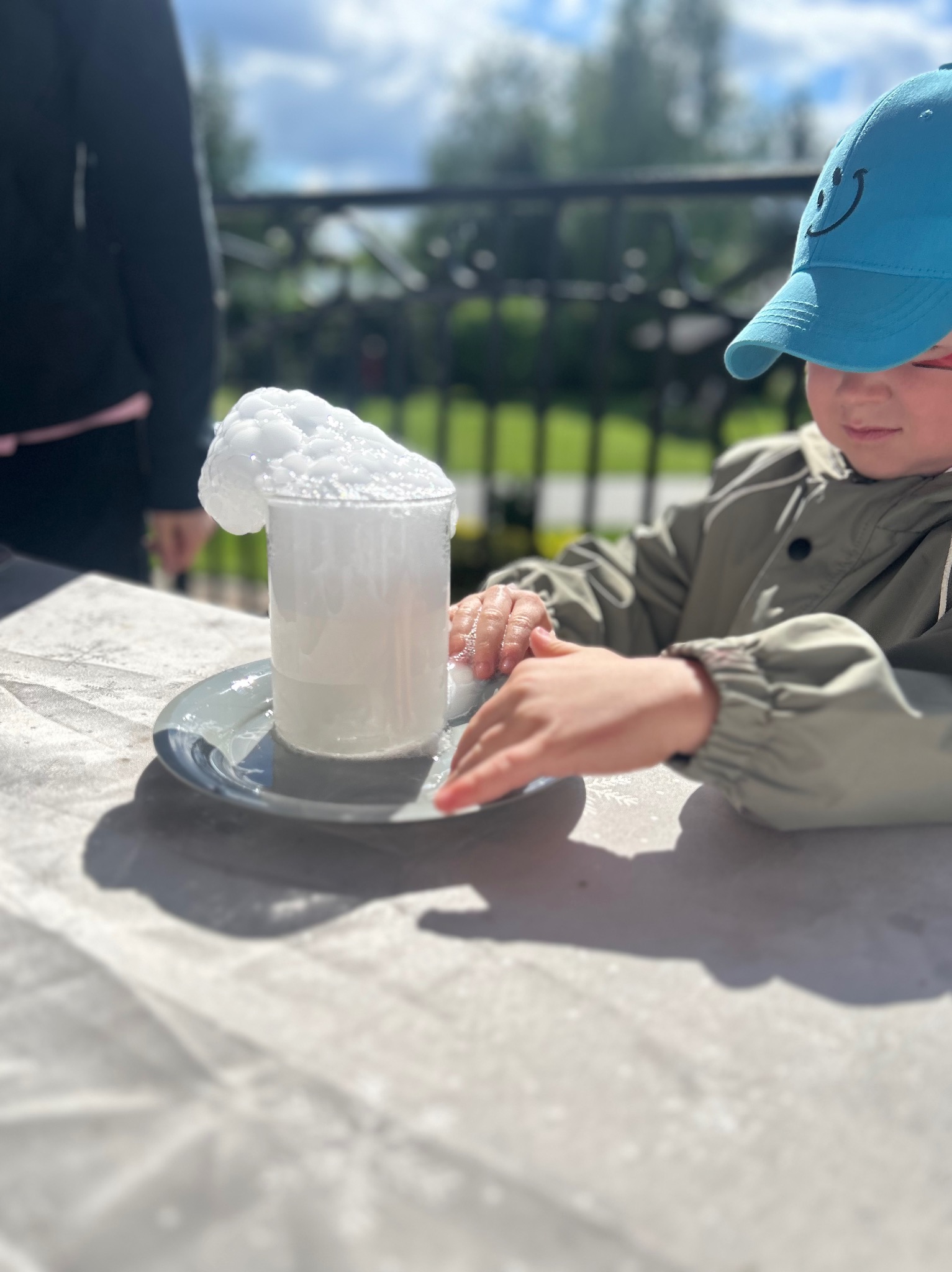 Chłopiec w czapce z uśmiechem eksperymentuje z cieczą i pianą w naczyniu na talerzu, na zewnątrz, w tle zieleń i balustrada.