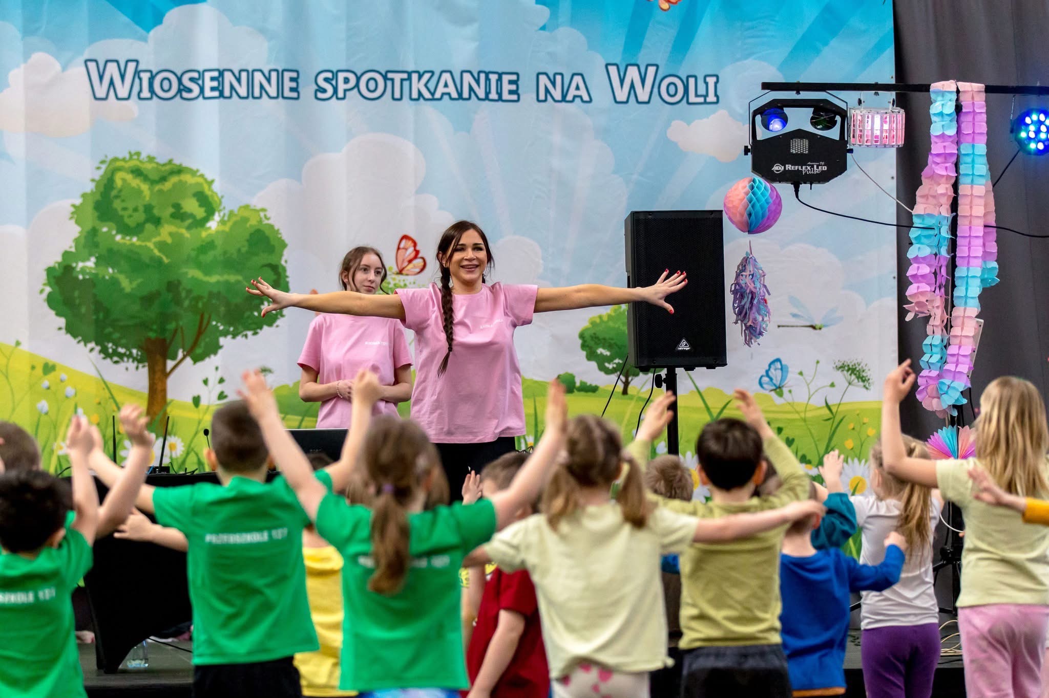 Animator prowadzi zabawę z dziećmi na tle sceny z napisem 'Wiosenne spotkanie na Woli', oświetlenie sceniczne i dekoracje.