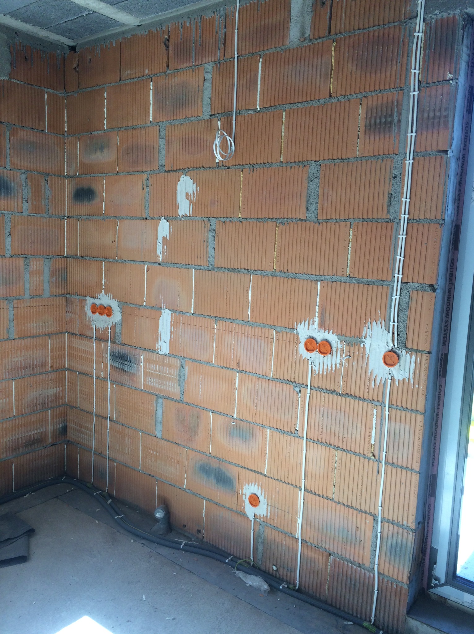 Instalacja elektryczna w surowym wnętrzu: pomarańczowe puszki pod gniazdka i białe kable prowadzone po ceglanej ścianie, widoczne przewody na podłodze.