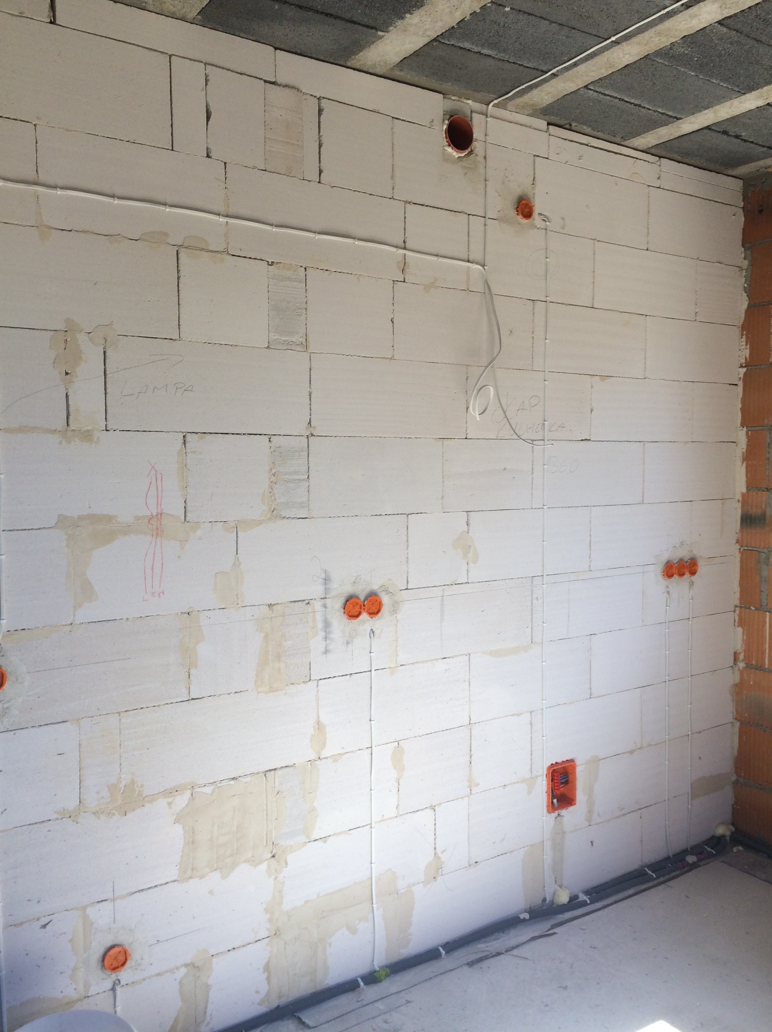 Ściana z betonu komórkowego w trakcie prac elektrycznych: widoczne kable, puszki instalacyjne, oznaczenia lamp. Surowy stan wykończenia wnętrza.