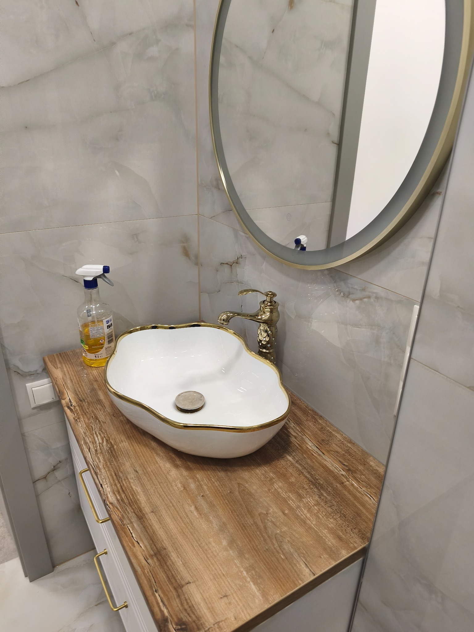 Umywalka w łazience z blatem imitującym drewno, złota armatura i okrągłe lustro. Na blacie stoi butelka ze środkiem czyszczącym. Ściany wyłożone płytkami imitującymi marmur.