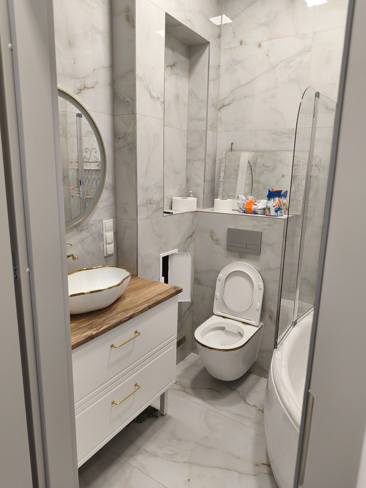 Elegancka łazienka z marmurowymi płytkami, białą umywalką na drewnianym blacie, złotymi akcentami i kabiną prysznicową. Widok na toaletę i lustro.