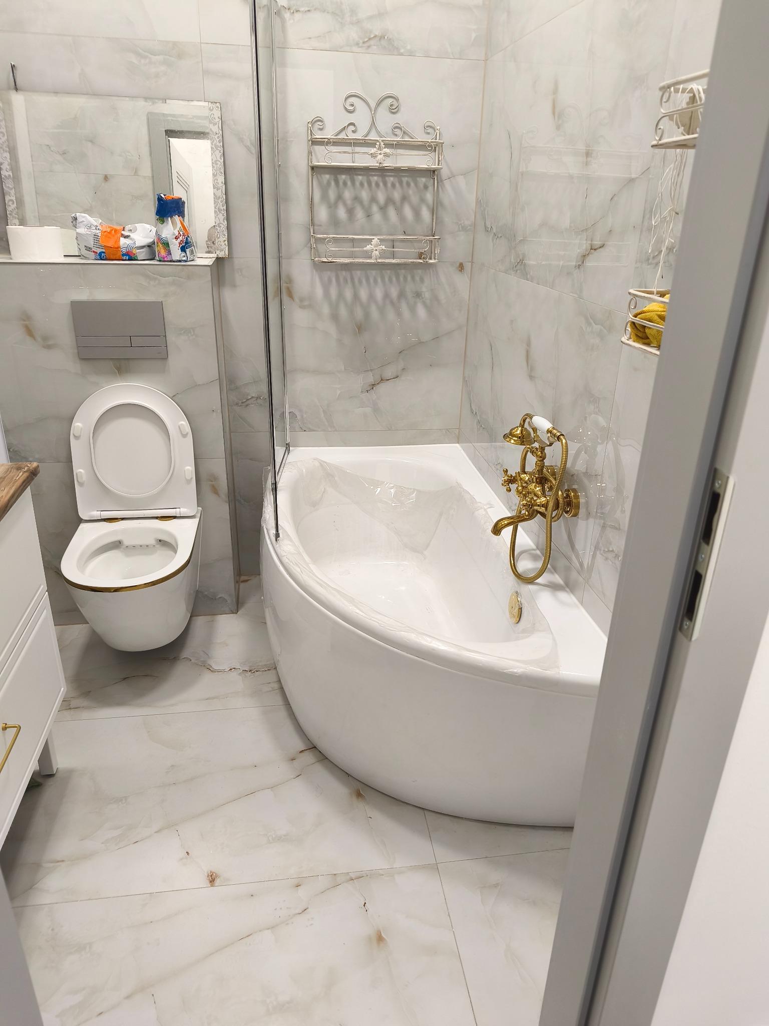 Elegancka łazienka z marmurowymi płytkami, białą wanną z złotą armaturą i toaletą. Widoczne półki dekoracyjne i lustro. Całość utrzymana w jasnej tonacji.