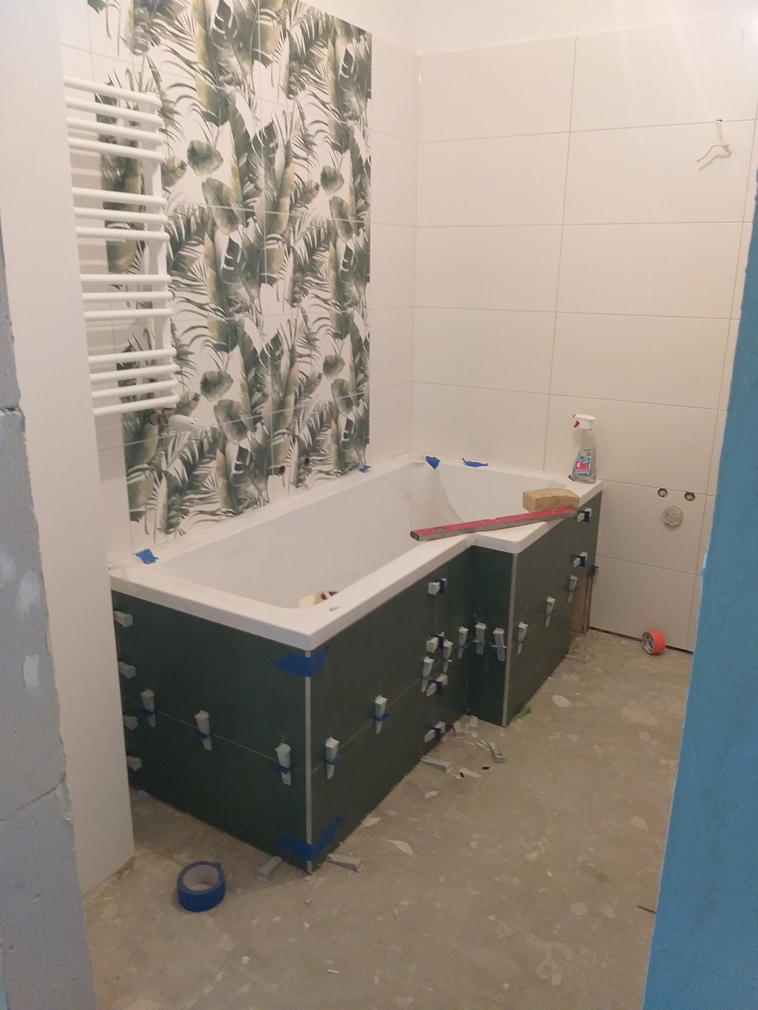 Wanna w trakcie obudowy z płytek, z widocznymi krzyżykami dystansowymi i poziomnicą. Na ścianie płytki z motywem roślinnym i grzejnik drabinkowy.