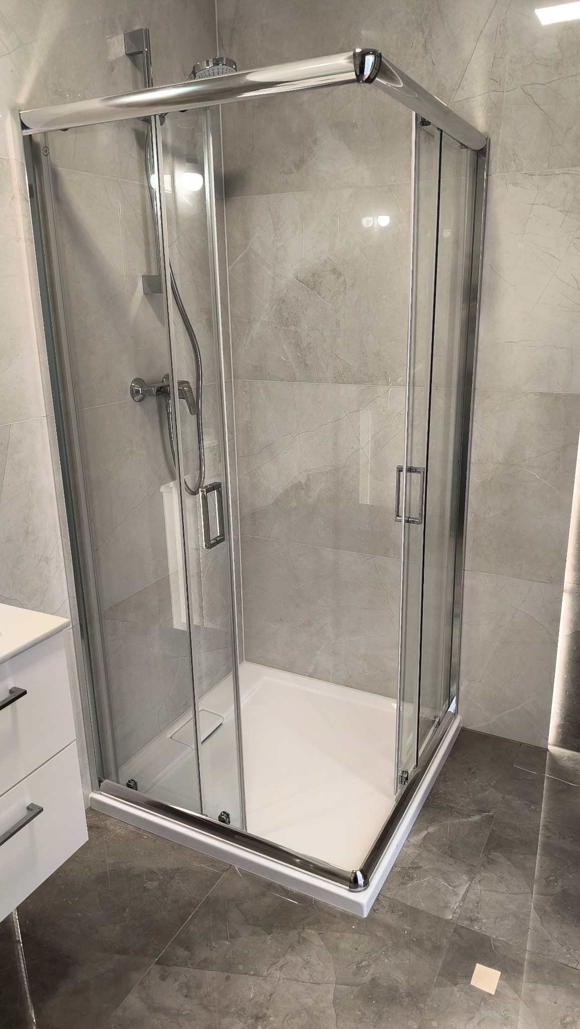 Nowoczesna kabina prysznicowa z chromowanymi profilami i szklanymi drzwiami, na tle szarych płytek imitujących marmur. Widoczny fragment szafki umywalkowej.