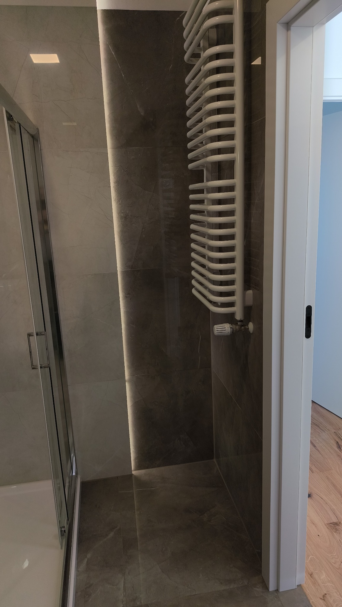 Wykończona łazienka z kabiną prysznicową, grzejnikiem drabinkowym i płytkami imitującymi marmur w odcieniach szarości. Widoczny fragment drewnianej podłogi za otwartymi drzwiami.