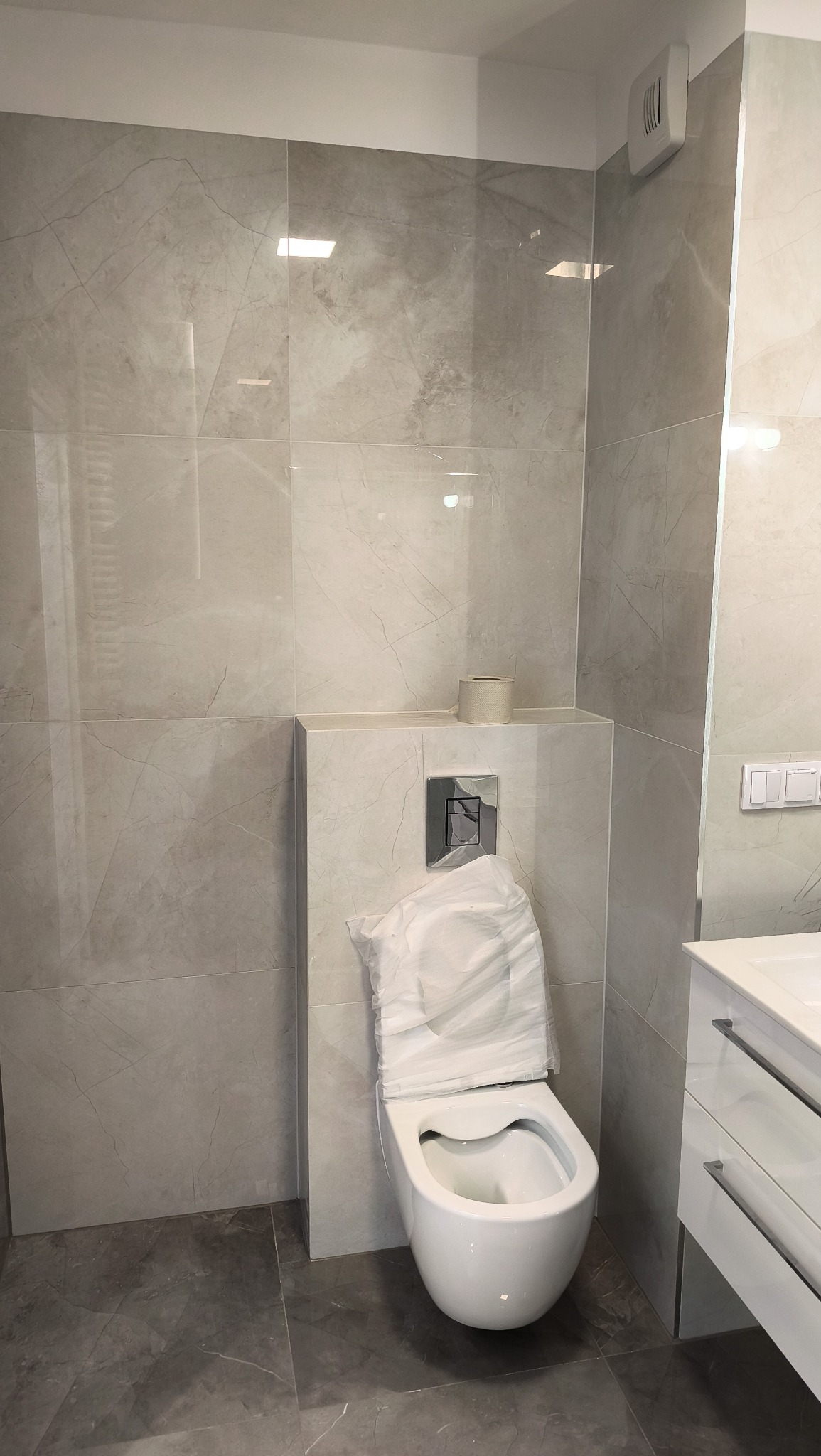 Nowoczesna łazienka w szarościach: marmurowe płytki, podwieszana toaleta z osłoną, biała szafka z umywalką. Minimalistyczny design, świeżo po remoncie.