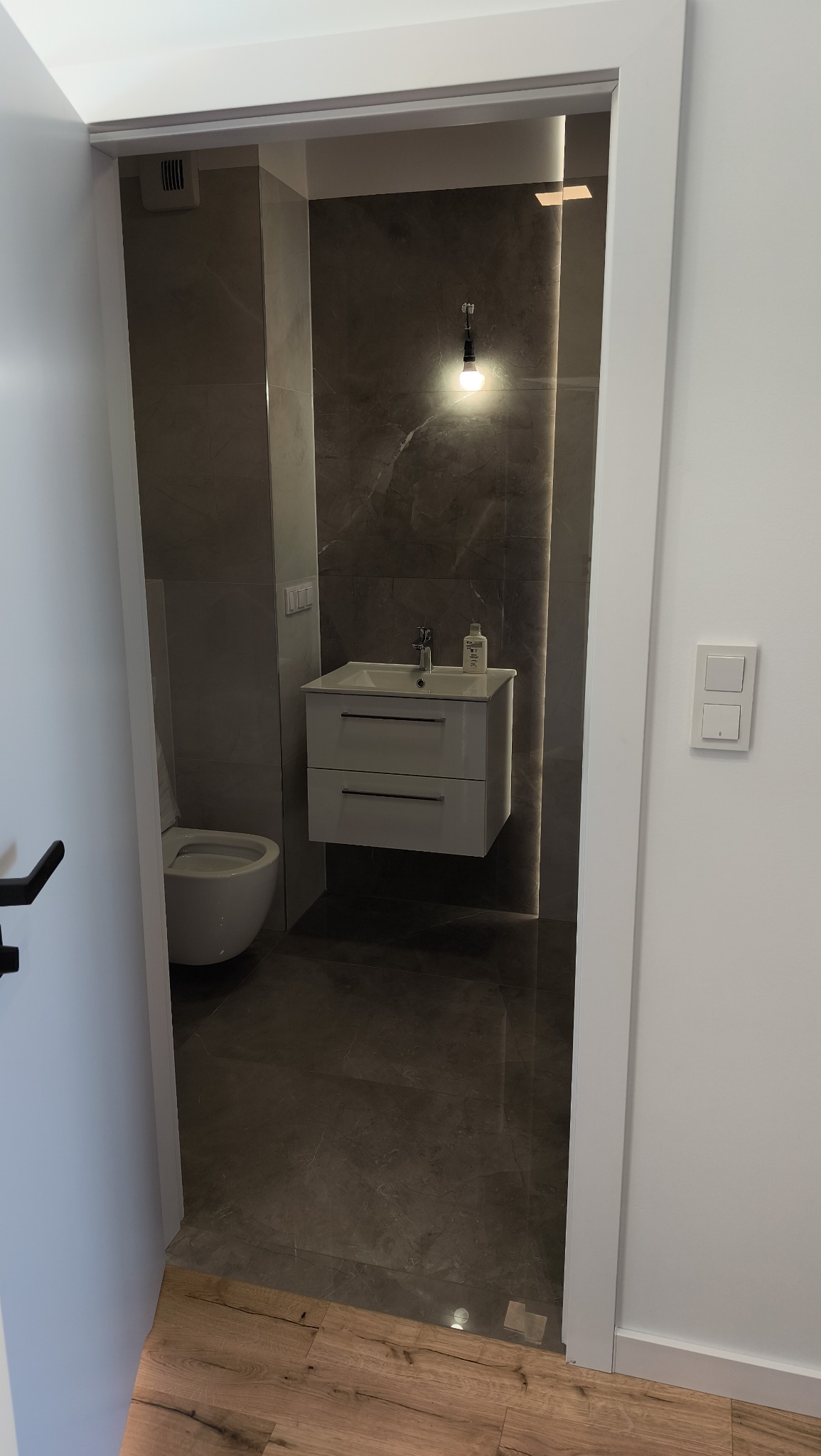 Nowoczesna łazienka z szarymi płytkami imitującymi marmur, podwieszaną szafką z umywalką i toaletą. Widok zza białych futryn drzwi, z akcentem na oświetlenie.