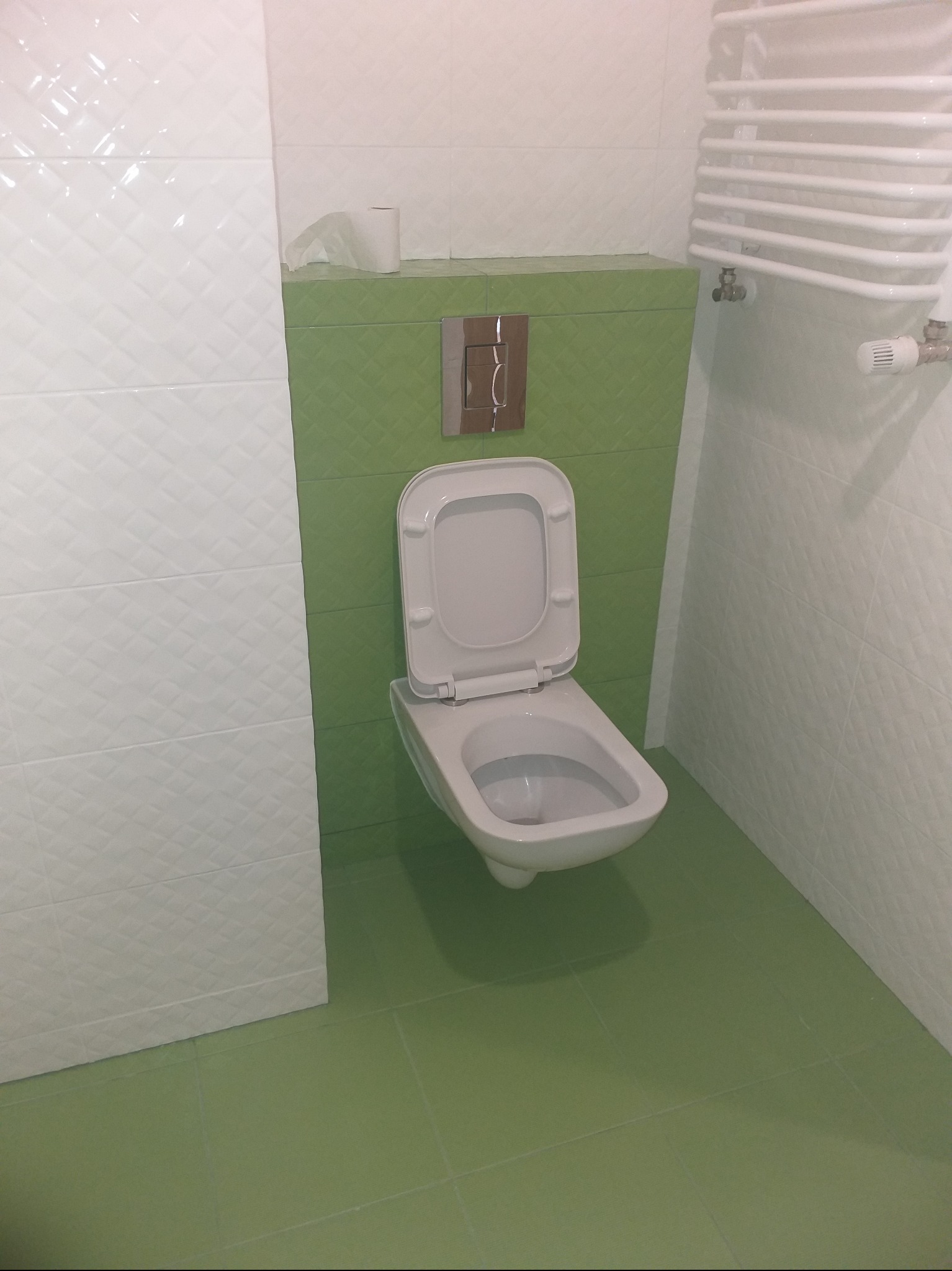 Nowoczesna toaleta podwieszana z otwartą klapą, zielone płytki na podłodze i ścianie za toaletą, białe płytki z wzorem na pozostałych ścianach, grzejnik drabinkowy.