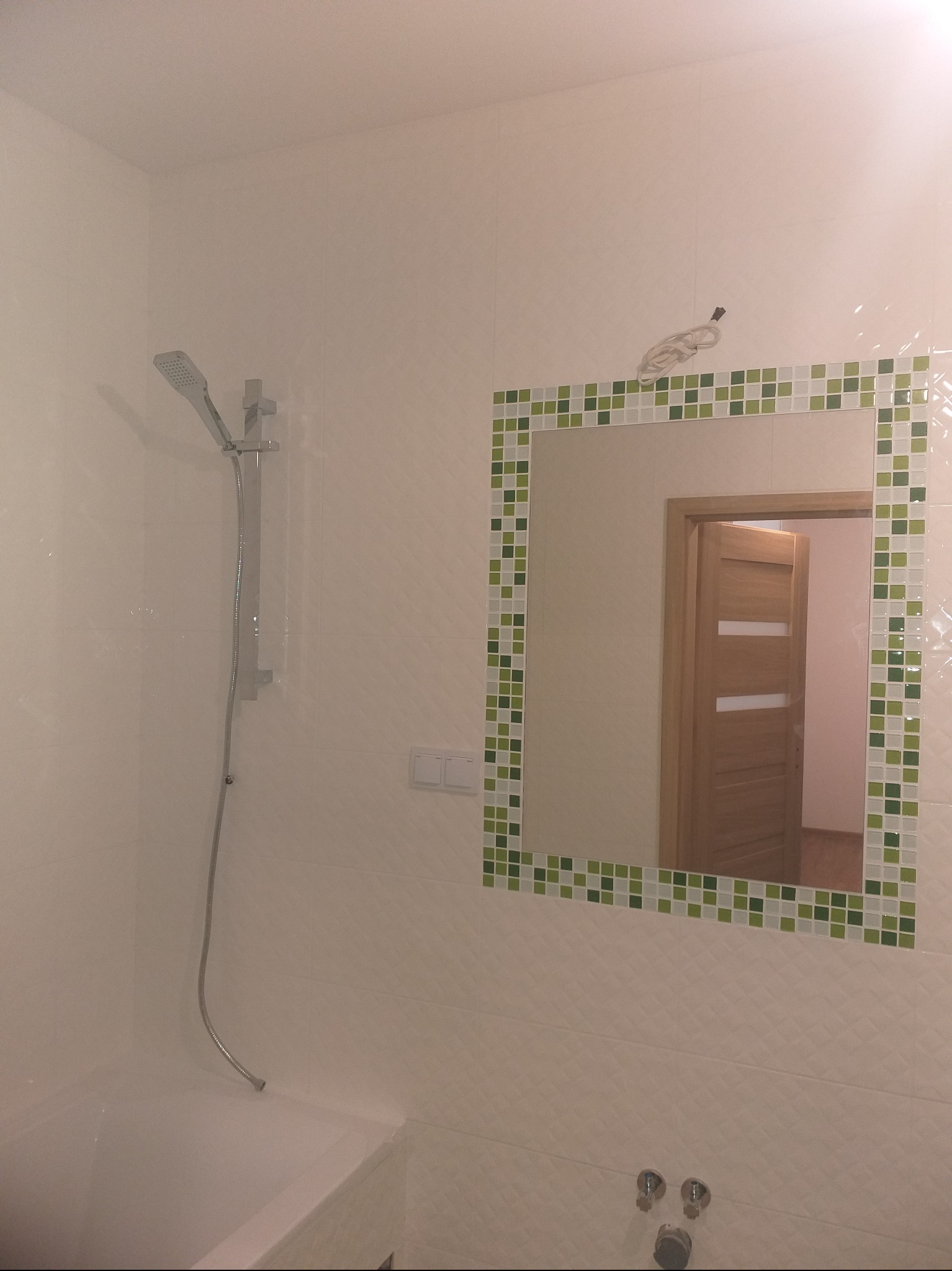 Wykończona łazienka z kabiną prysznicową, lustrem w ramie z mozaiki i widoczną w odbiciu drewnianą ościeżnicą drzwi. Białe płytki na ścianach.