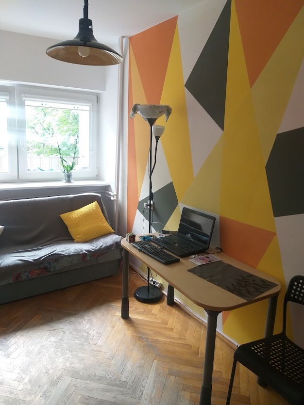 Pomysłowe malowanie ścian w geometryczne wzory w odcieniach żółci, pomarańczy i szarości, tworzące unikalną dekorację pokoju z biurkiem i kanapą.