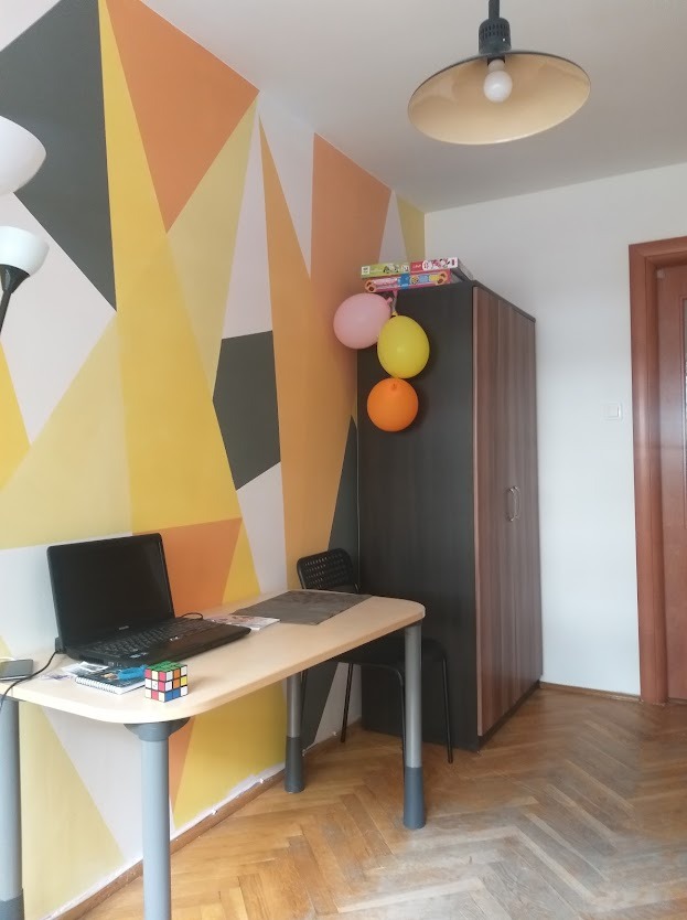 Pokój z geometrycznym malowaniem ścian w odcieniach żółci i szarości. Biurko z laptopem, szafa z balonami, drewniana podłoga. Nietypowy wzór na ścianie.