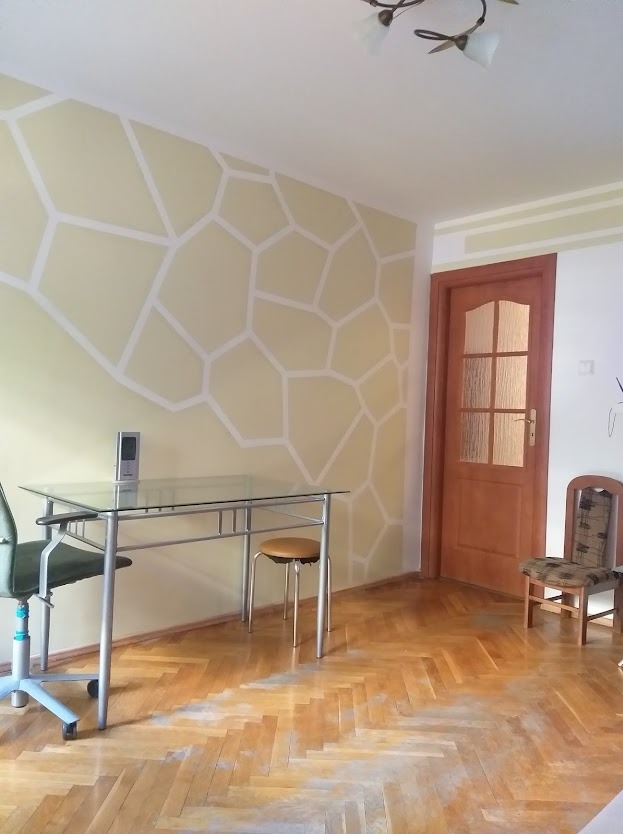 Pokój z wzorem geometrycznym na ścianie w odcieniach beżu i bieli. Widoczny stół ze szklanym blatem, krzesło biurowe i drewniana podłoga w jodełkę.