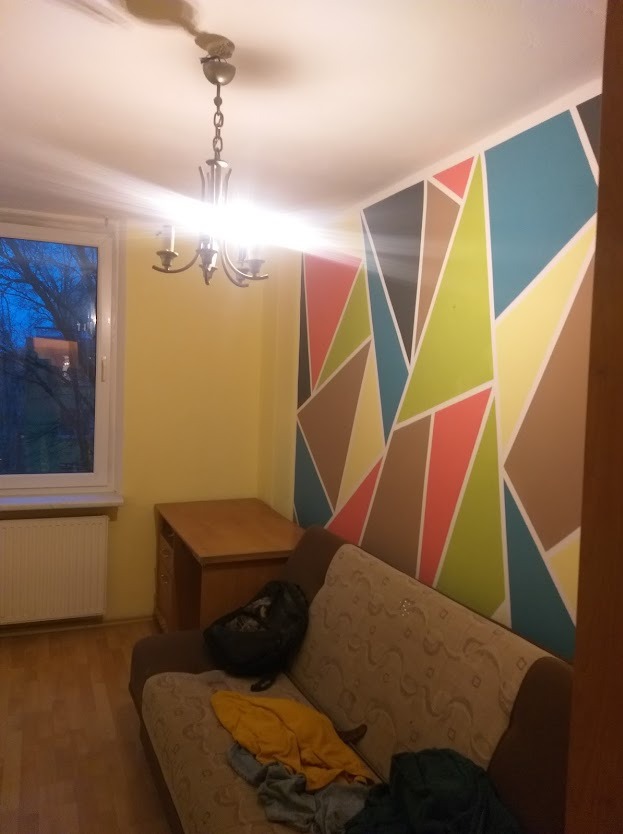 Pokój z geometrycznym malowaniem ściany w odcieniach brązu, zieleni, błękitu i żółci. Obok stoi biurko i sofa. W oknie widoczne drzewa. Całość oświetla żyrandol.