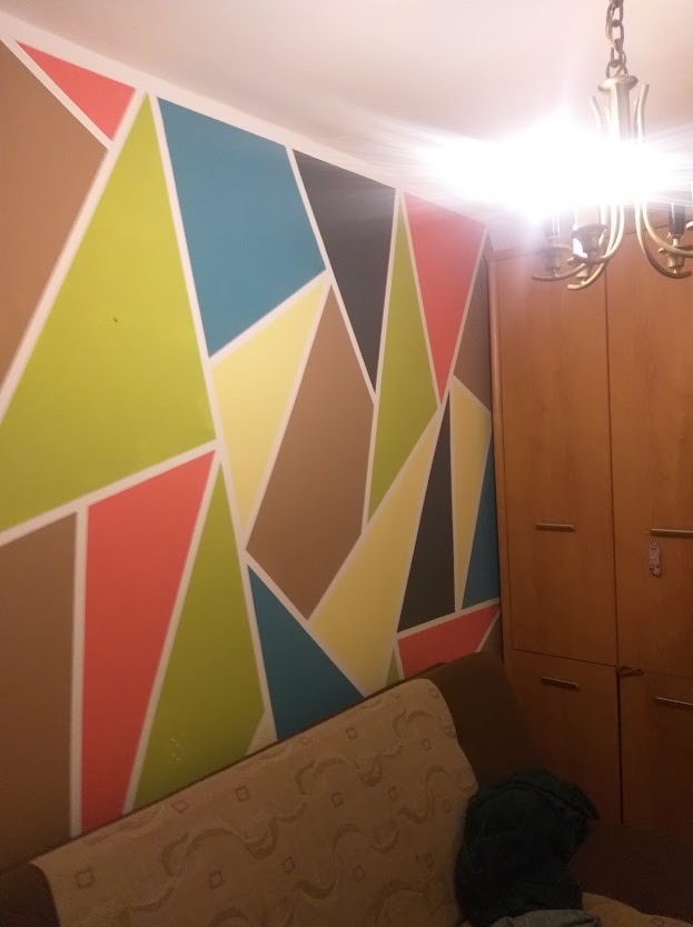 Wzorzysta ściana w pokoju z geometrycznym malowaniem w różnych kolorach, oddzielona białymi liniami. Częściowo widoczny beżowy narożnik i drewniana szafa.
