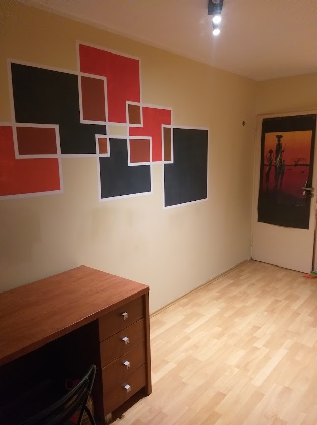 Pokój z geometrycznym malowaniem ścian w odcieniach czerwieni i czerni, obok biurko z szufladami i drzwi z afrykańskim motywem. Nietypowa dekoracja wnętrza.