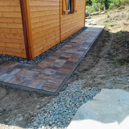 Marcin Stachura - Ścieżka z kostki brukowej prowadząca do drewnianego domku, obrzeża wysypane drobnymi kamieniami. Ujęcie z boku budynku, widoczne okno z okiennicą.