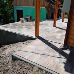 Marcin Stachura - Ułożona kostka brukowa na tarasie z drewnianą konstrukcją, widoczne wiaderka z farbą i fragment budynku w tle. Kostka w odcieniach szarości i brązu, świeżo ułożona.