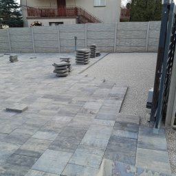 Marcin Stachura - Układanie szarej kostki brukowej na posesji z widocznym ogrodzeniem panelowym i stertami kostki. Częściowo wyłożona powierzchnia, przygotowana pod dalsze prace.