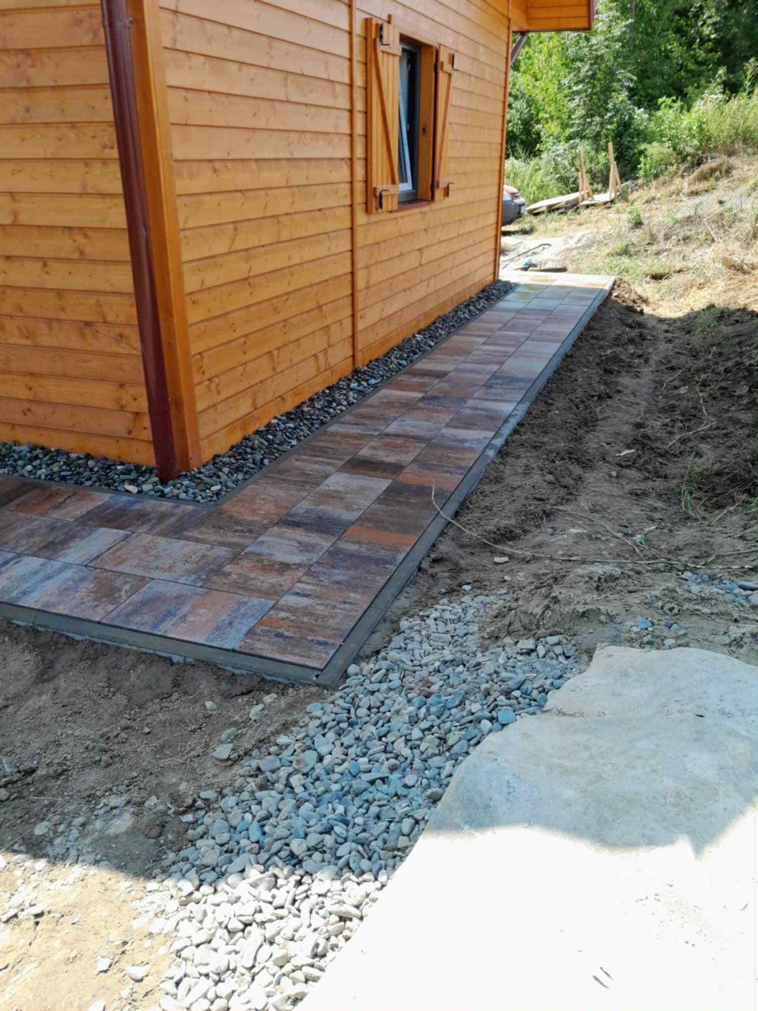 Ułożona ścieżka z kostki brukowej prowadząca wzdłuż drewnianego domku, obłożona drobnymi kamieniami. Widoczne nierówności terenu i fragment zieleni w tle.