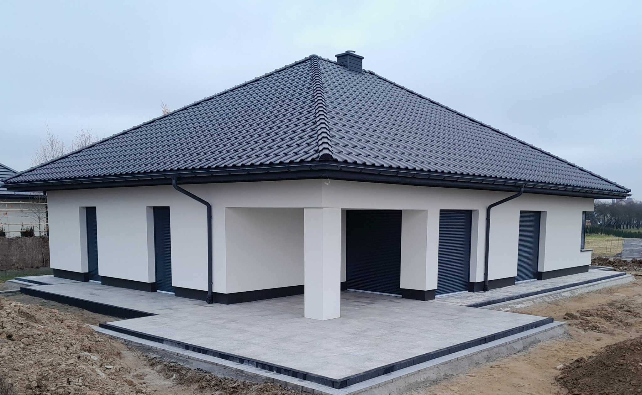 Nowoczesny dom parterowy z szarą dachówką i jasną elewacją. Wykończony taras z kostki brukowej. Widoczne przygotowania do zagospodarowania terenu wokół domu.