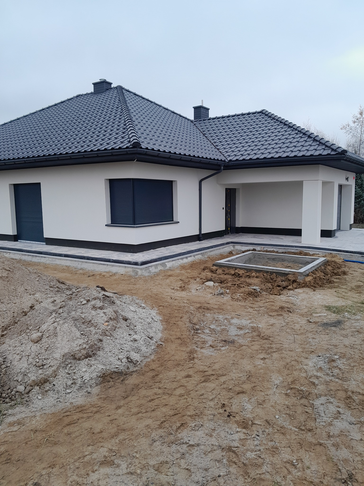 Nowoczesny dom jednorodzinny z kostką brukową i przygotowanym wykopem pod szambo w Dębicy. Widoczny fragment ogrodu i elewacja budynku.