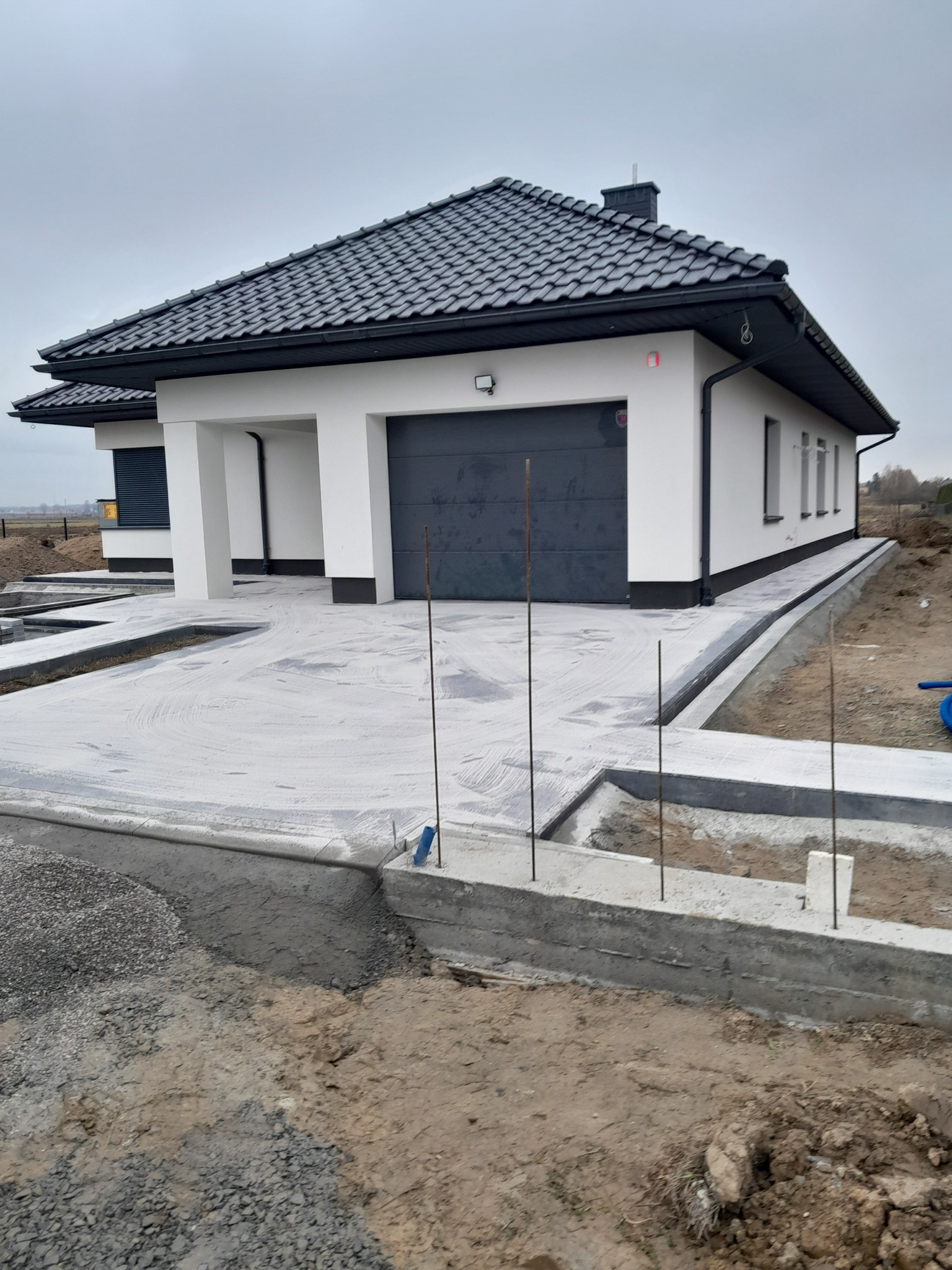 Nowoczesny dom jednorodzinny z szarą bramą garażową i przygotowanym podjazdem z podsypką pod kostkę brukową. Widoczne obrzeża i teren wokół w trakcie prac.