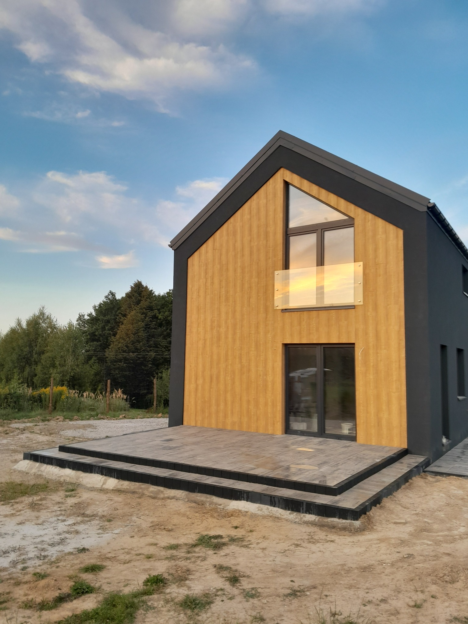 Nowoczesny dom z drewnianą elewacją i tarasem z kostki brukowej, widok z dołu na tle błękitnego nieba i zieleni. Wykończony taras z kostki brukowej.