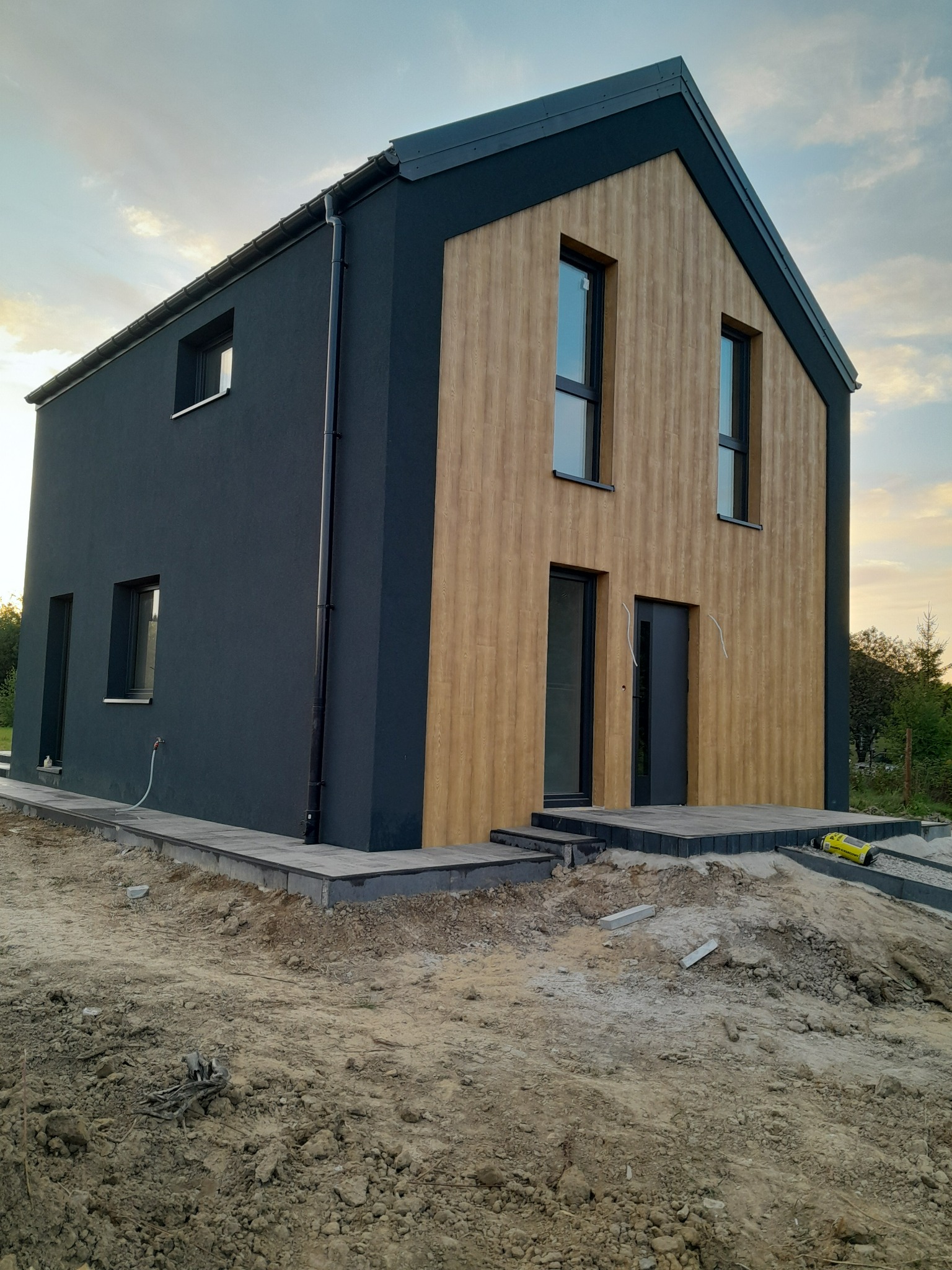 Nowoczesny dom z elewacją w kolorze grafitu i drewna, z kostką brukową wokół. Widoczne przygotowania terenu i narzędzia. Ujęcie z dołu, podkreślające bryłę budynku.