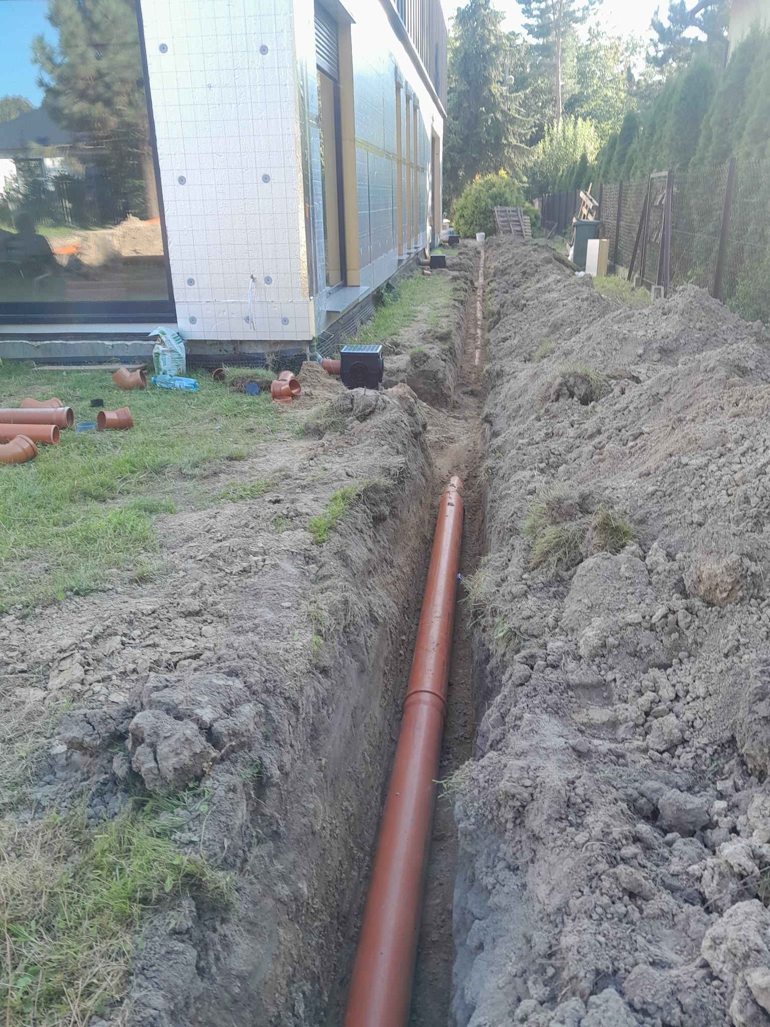 Brązowa rura ułożona w wykopie ziemnym wzdłuż budynku z ociepleniem. Na trawie leżą dodatkowe rury i akcesoria. W tle widoczne drzewa i ogrodzenie.