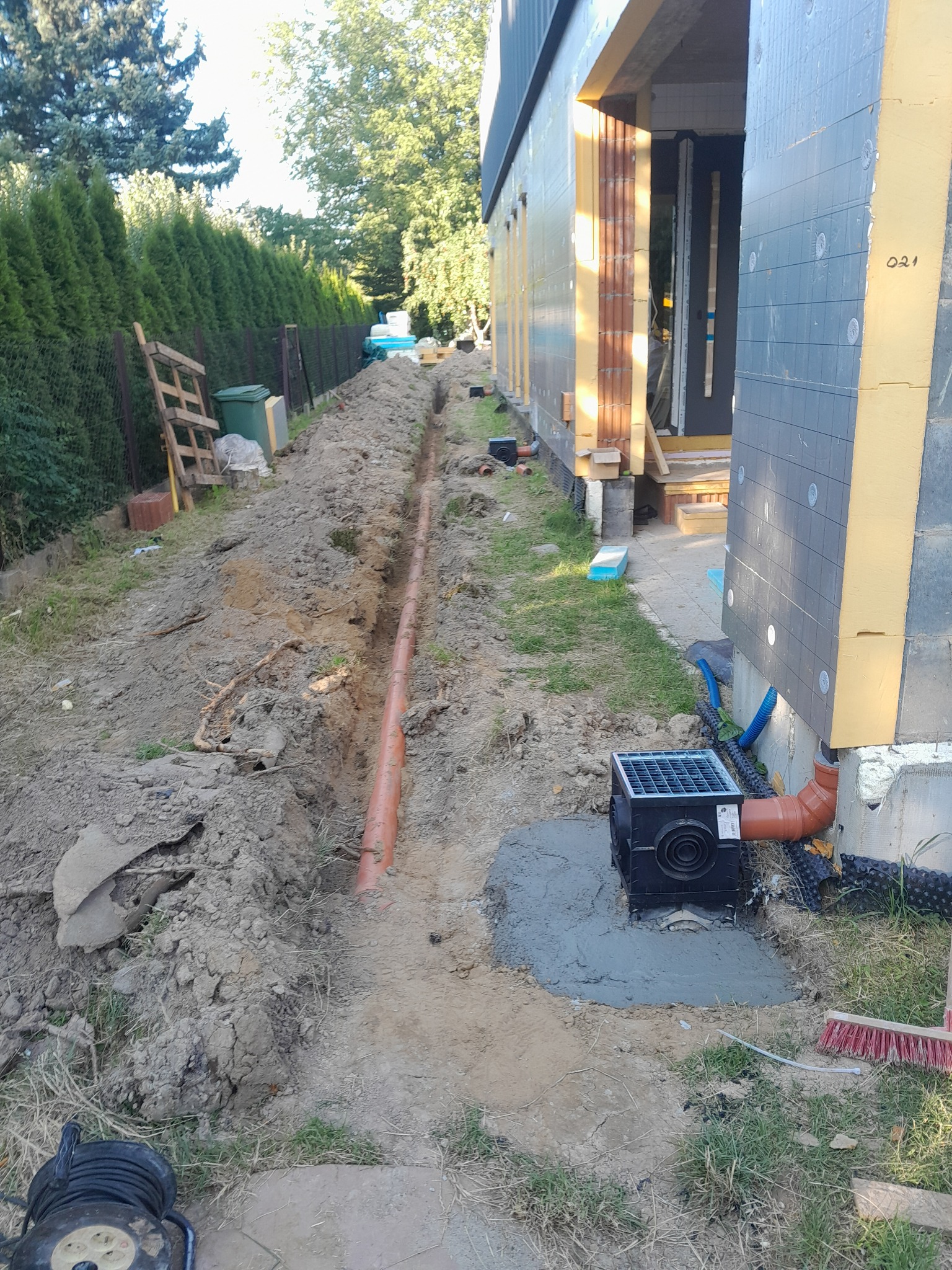 Wykopany rów z rurą odprowadzającą wodę deszczową do studzienki przy budynku z ociepleniem. Widoczne narzędzia i elementy instalacji.